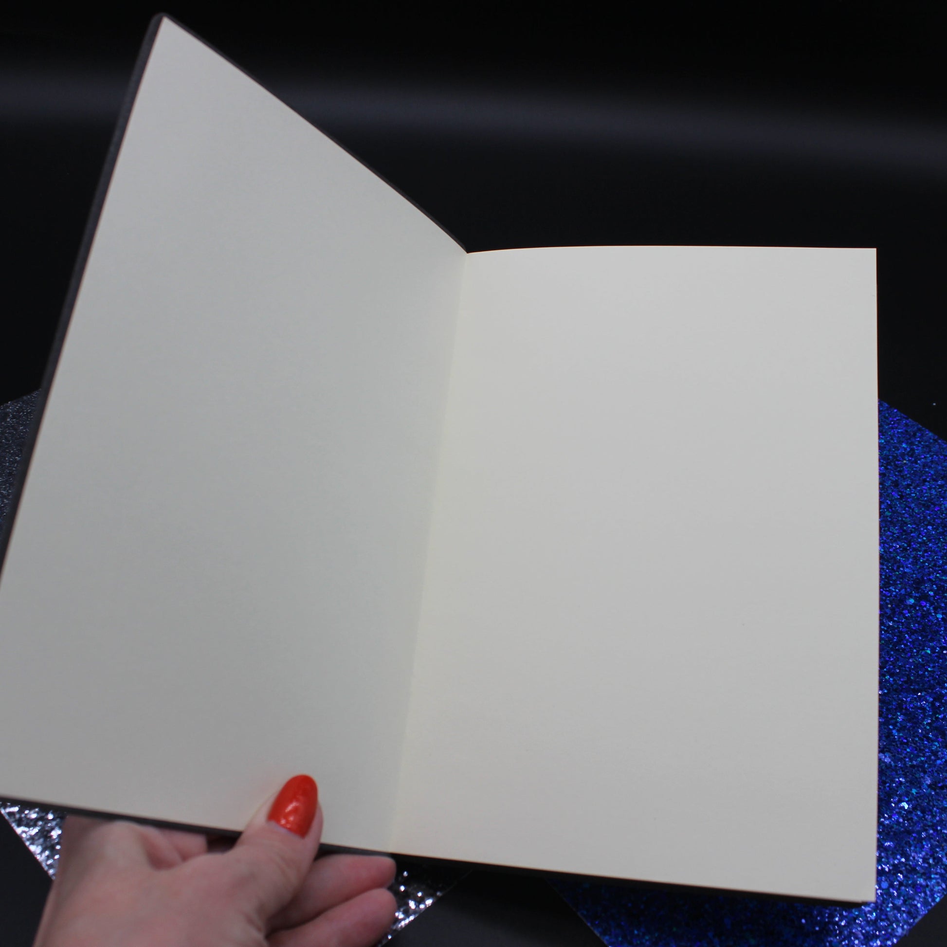 An open blank notebook or card album with cream-colored pages, held by a hand with red nail polish over a black background with glittery blue and silver paper sheets.