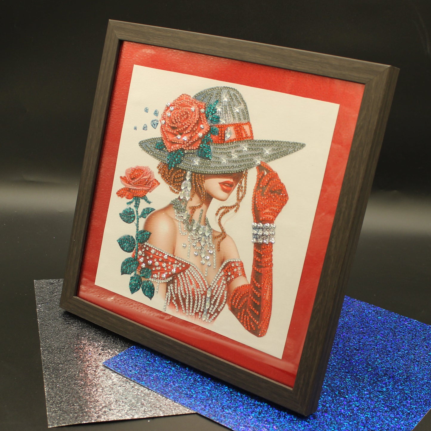 A framed art print featuring a stylized illustration of a woman in a wide-brimmed hat with red roses, displayed on a dark surface with glittery blue and silver sheets.