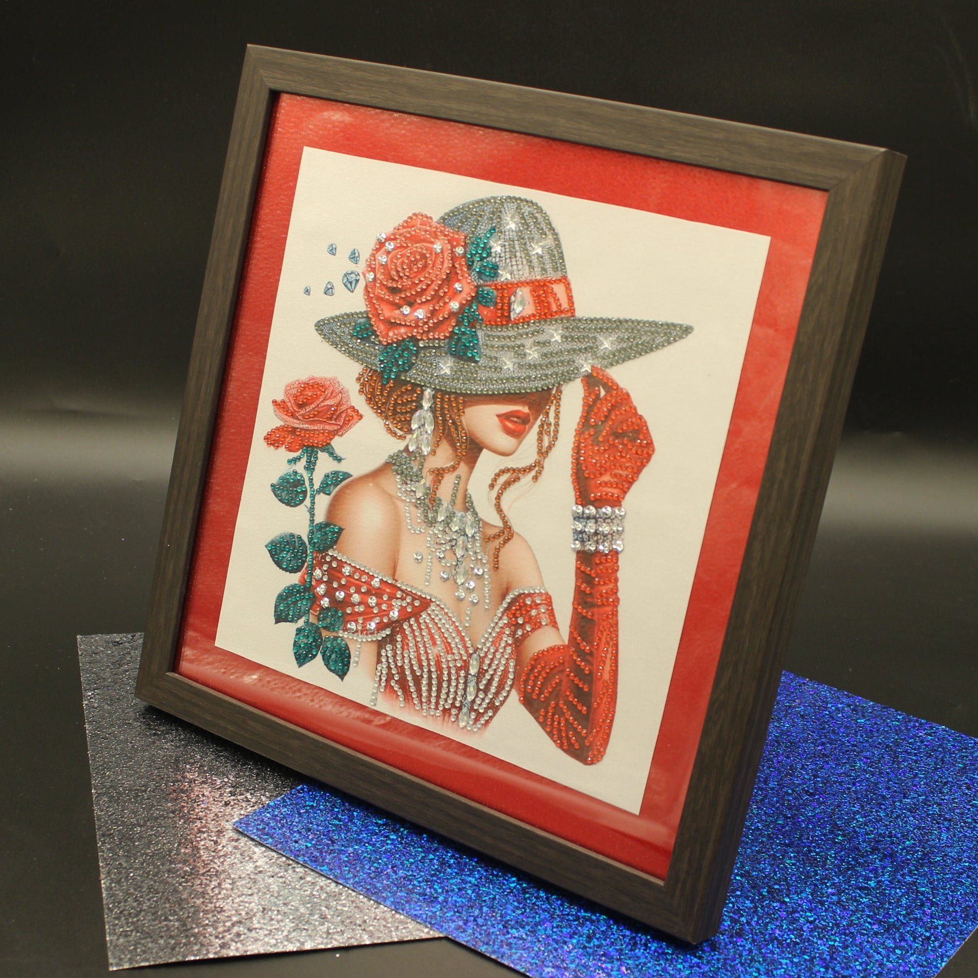A framed art print featuring a stylized illustration of a woman in a wide-brimmed hat with red roses, displayed on a dark surface with glittery blue and silver sheets.